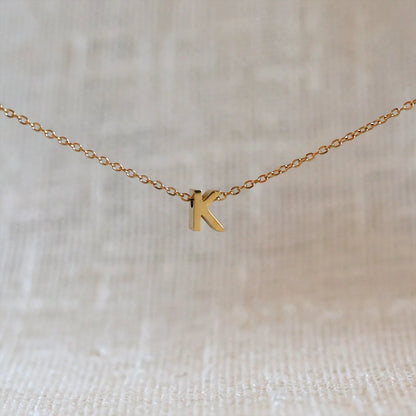 Collier L'instant avec pendentif lettre K doré en acier inoxydable, suspendu au centre d'une maille fine sur fond beige.