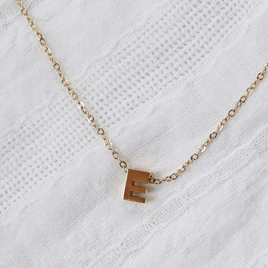 Zoom sur le collier L'instant, pendentif lettre E en acier inoxydable doré, capturé sur tissu beige texturé.