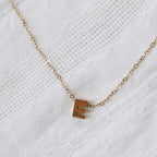 Zoom sur le collier L'instant, pendentif lettre E en acier inoxydable doré, capturé sur tissu beige texturé.