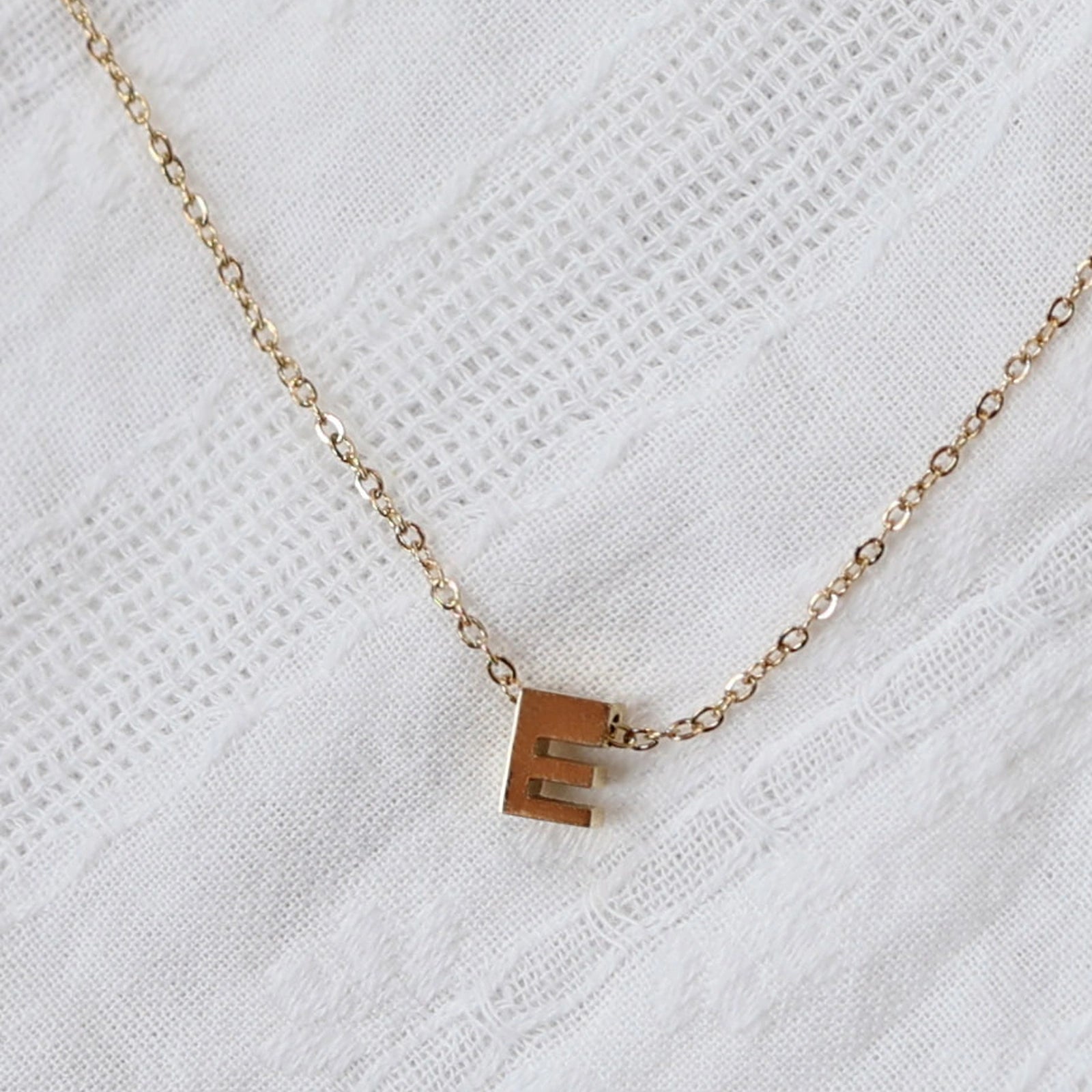 Zoom sur le collier L'instant, pendentif lettre E en acier inoxydable doré, capturé sur tissu beige texturé.