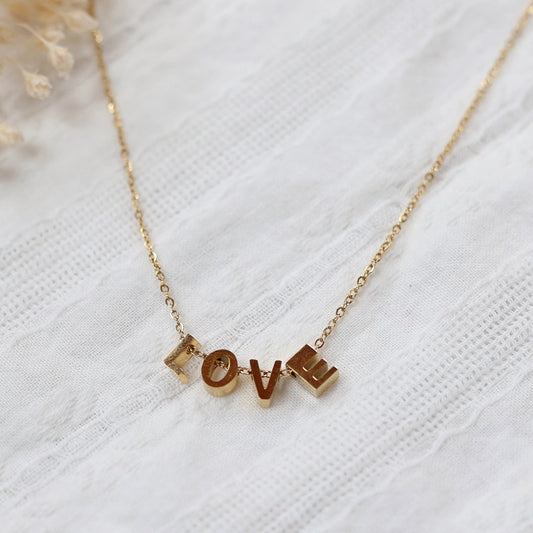 Collier L'instant avec pendentifs lettres LOVE en acier inoxydable doré, bijou délicat posé sur un tissu beige texturé.