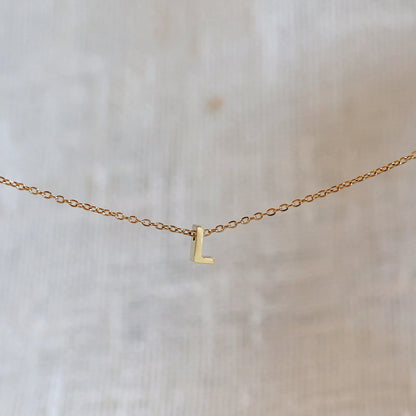 Collier L'instant avec pendentif lettre L doré en acier inoxydable, suspendu au centre d'une maille fine sur fond beige.