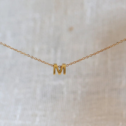 Collier L'instant avec pendentif lettre M doré en acier inoxydable, suspendu au centre d'une maille fine sur fond beige.