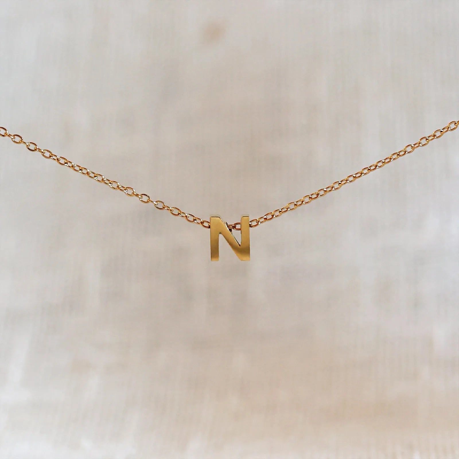 Collier L'instant avec pendentif lettre N doré en acier inoxydable, suspendu au centre d'une maille fine sur fond beige.
