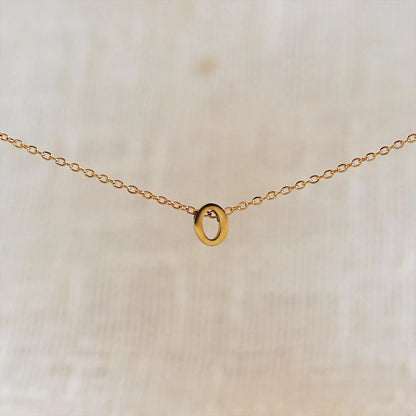 Collier L'instant avec pendentif lettre O doré en acier inoxydable, suspendu au centre d'une maille fine sur fond beige.