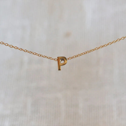 Collier L'instant avec pendentif lettre P doré en acier inoxydable, suspendu au centre d'une maille fine sur fond beige.