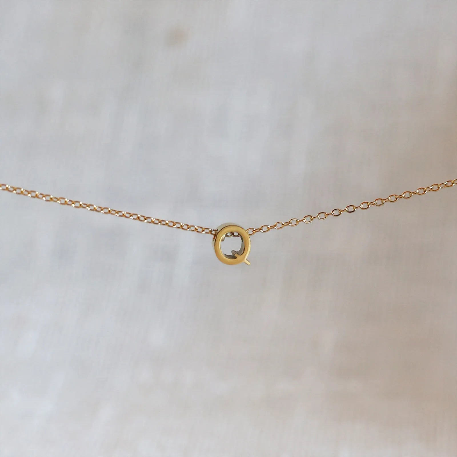 Collier L'instant avec pendentif lettre Q doré en acier inoxydable, suspendu au centre d'une maille fine sur fond beige.