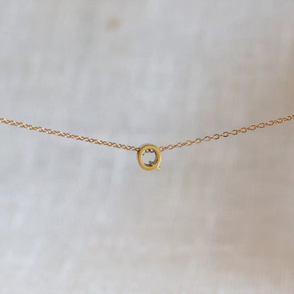 Collier L'instant avec pendentif lettre Q doré en acier inoxydable, suspendu au centre d'une maille fine sur fond beige.