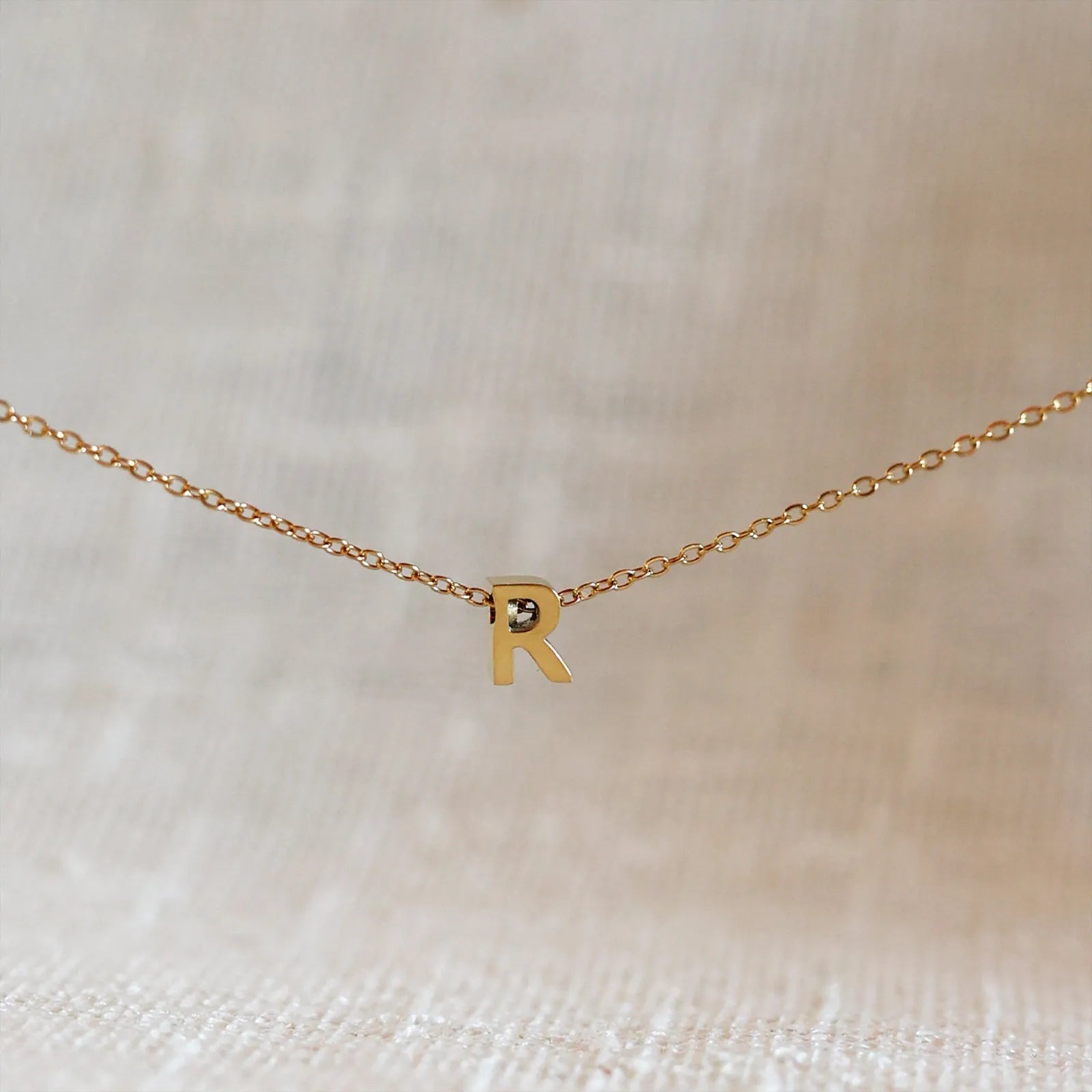 Collier L'instant avec pendentif lettre R doré en acier inoxydable, suspendu au centre d'une maille fine sur fond beige.