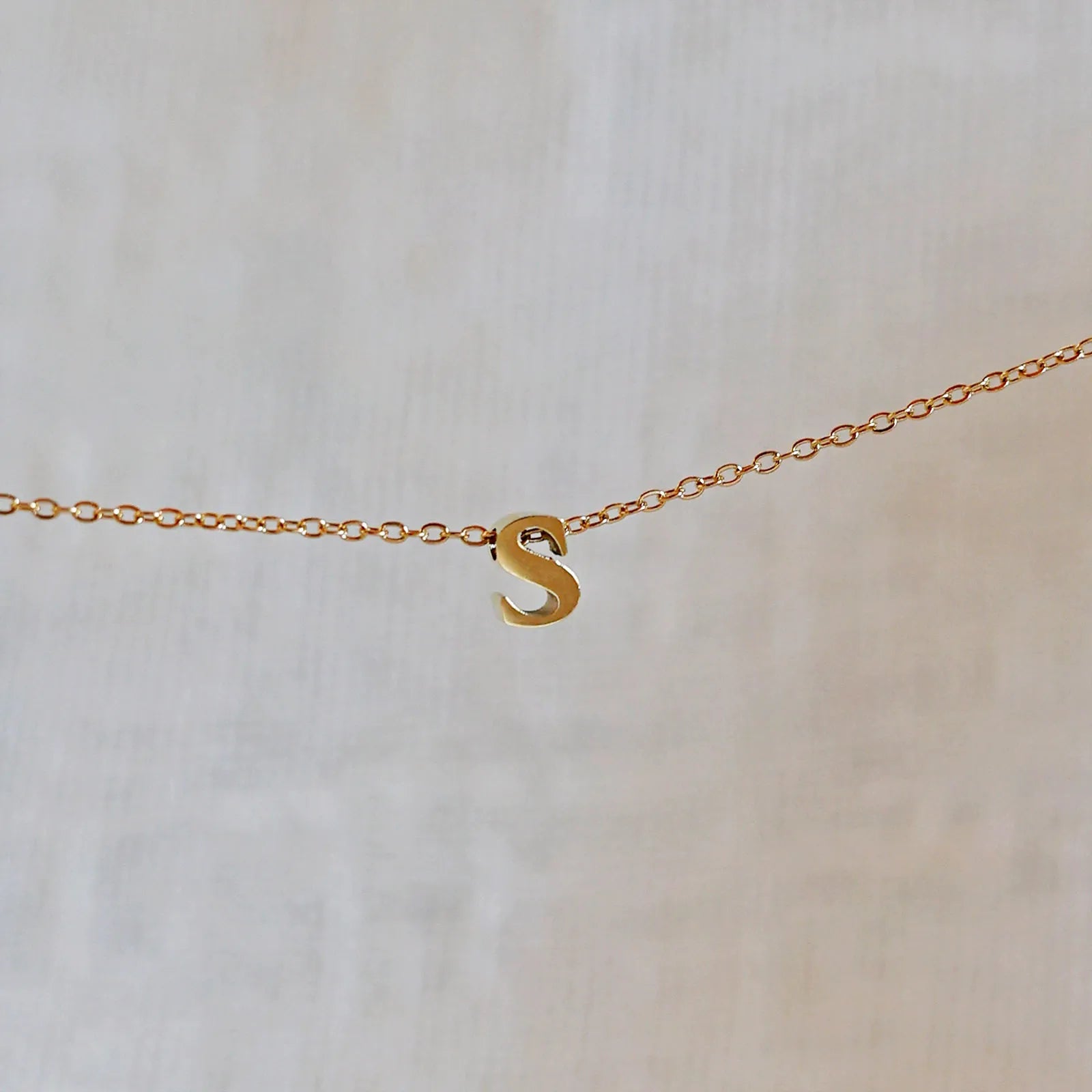 Collier L'instant avec pendentif lettre S doré en acier inoxydable, suspendu au centre d'une maille fine sur fond beige.