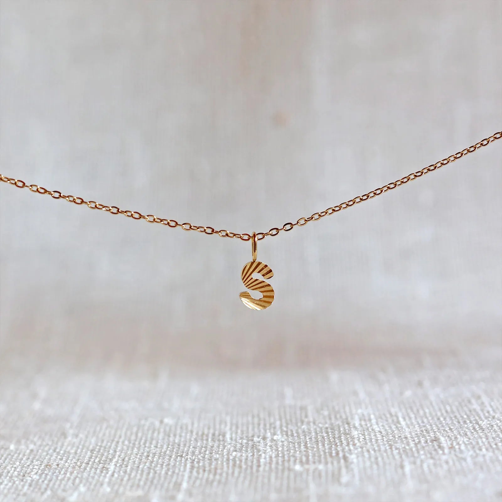 Collier "Je suis" avec pendentif lettre S striée en acier doré, suspendu sur un fond en tissu beige texturé.