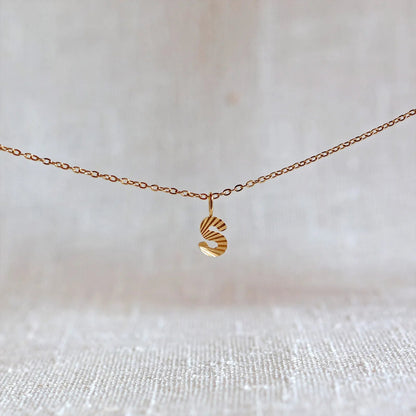 Collier "Je suis" avec pendentif lettre S striée en acier doré, suspendu sur un fond en tissu beige texturé.