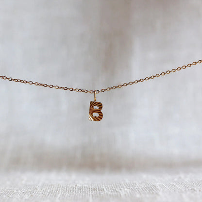 Collier "Je suis" avec pendentif lettre B striée en acier doré, suspendu sur un fond en tissu beige texturé.