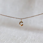 Collier "Je suis" avec pendentif lettre C striée en acier doré, suspendu sur un fond en tissu beige texturé.