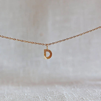 Collier "Je suis" avec pendentif lettre D striée en acier doré, suspendu sur un fond en tissu beige texturé.