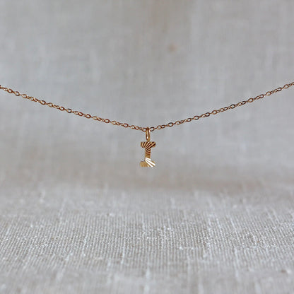 Collier "Je suis" avec pendentif lettre I striée en acier doré, suspendu sur un fond en tissu beige texturé.