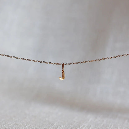 Collier "Je suis" avec pendentif lettre J striée en acier doré, suspendu sur un fond en tissu beige texturé.