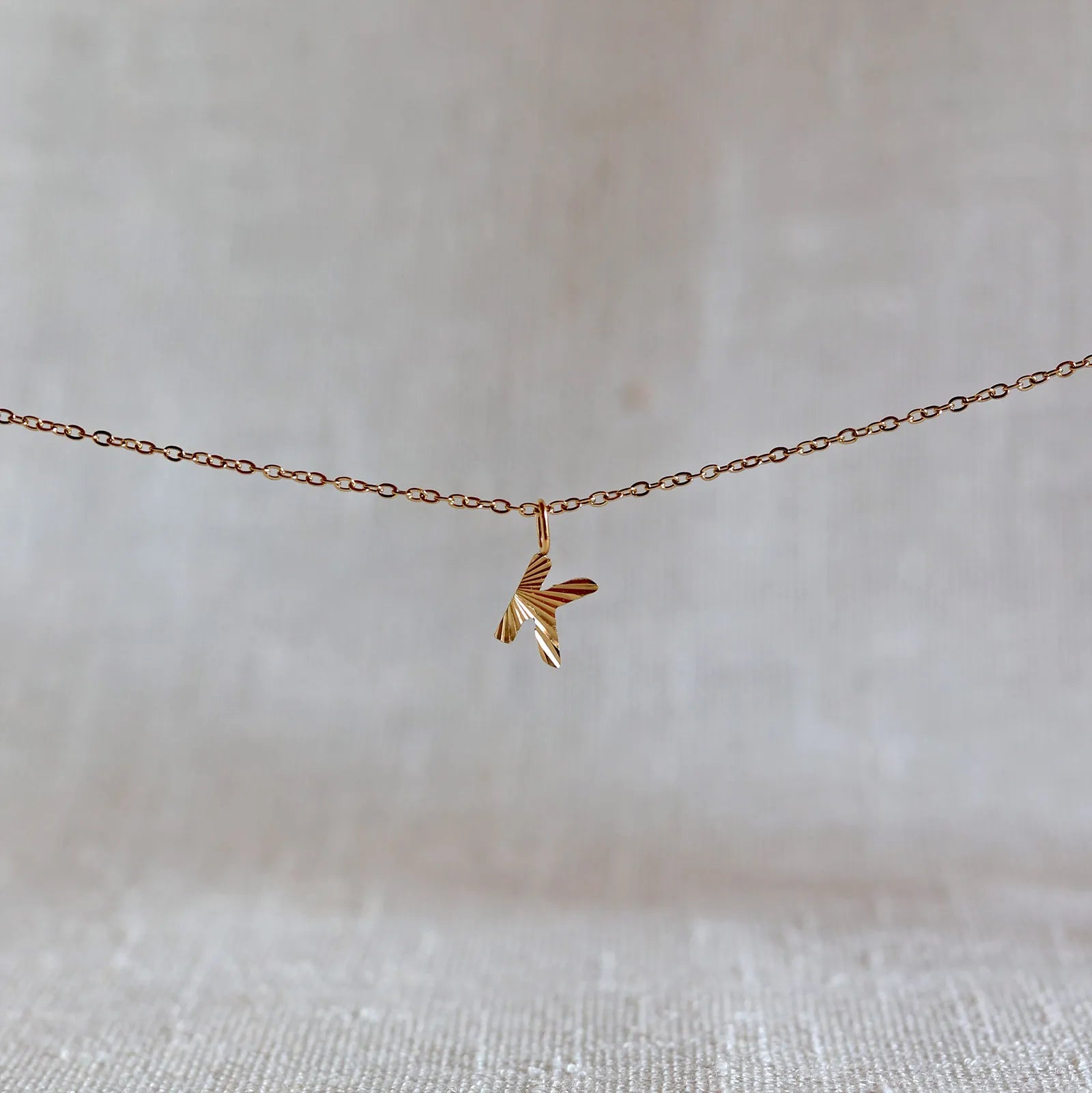 Collier "Je suis" avec pendentif lettre K striée en acier doré, suspendu sur un fond en tissu beige texturé.
