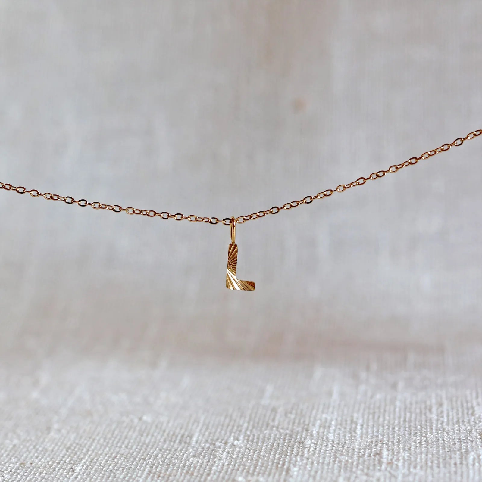 Collier "Je suis" avec pendentif lettre L striée en acier doré, suspendu sur un fond en tissu beige texturé.