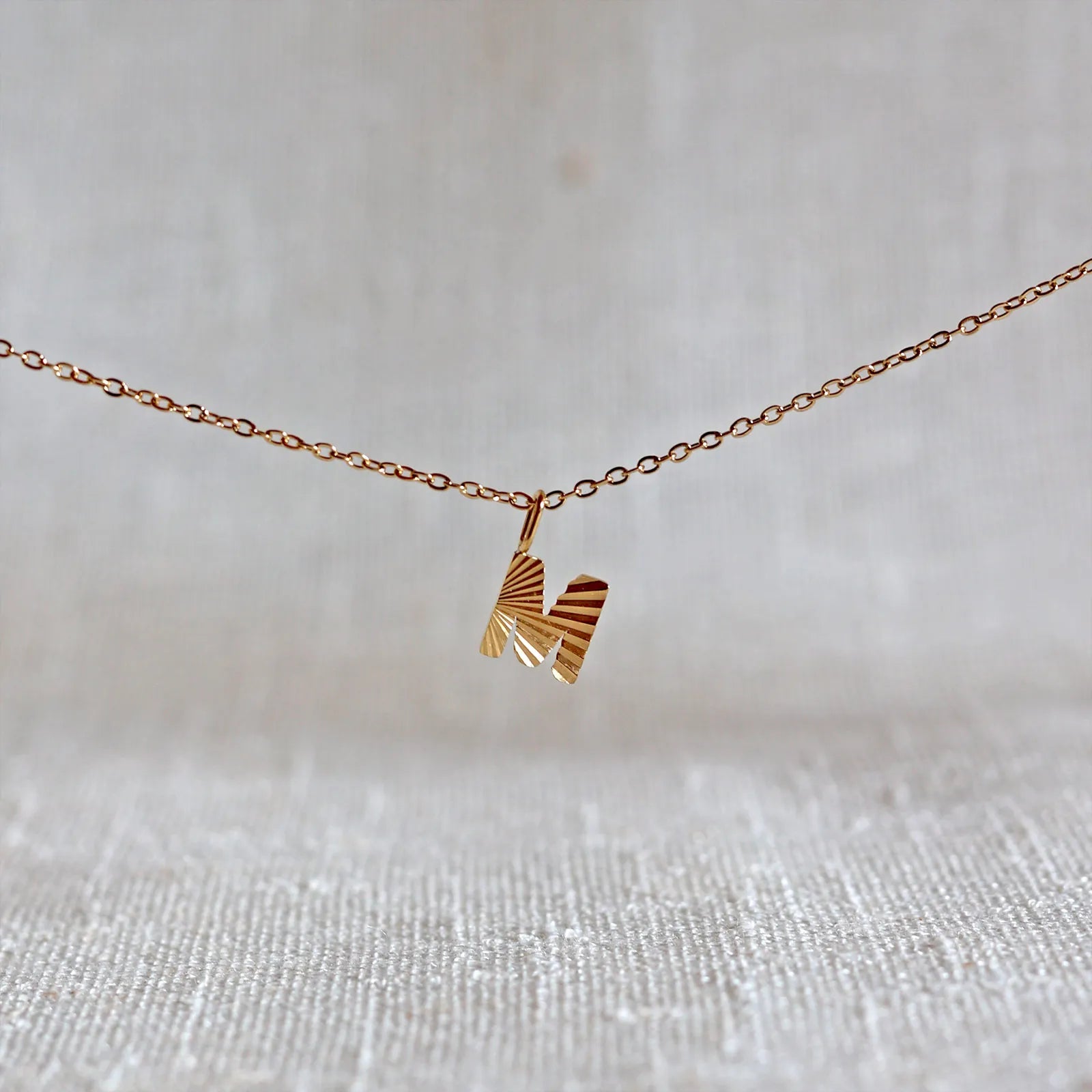 Collier "Je suis" avec pendentif lettre M striée en acier doré, suspendu sur un fond en tissu beige texturé.