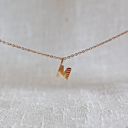 Collier "Je suis" avec pendentif lettre M striée en acier doré, suspendu sur un fond en tissu beige texturé.