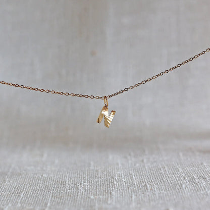 Collier "Je suis" avec pendentif lettre N striée en acier doré, suspendu sur un fond en tissu beige texturé.