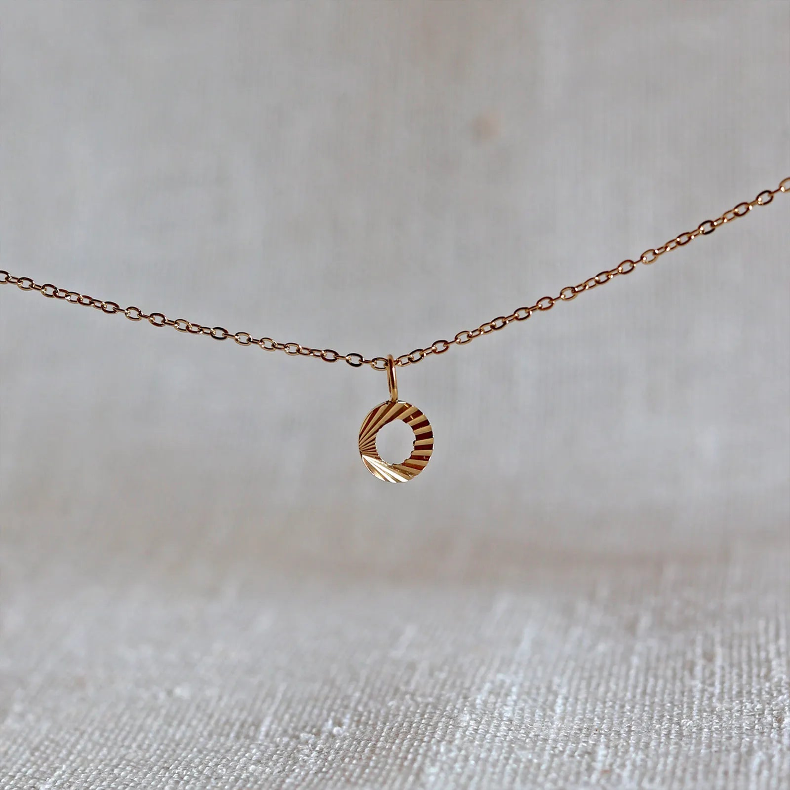 Collier "Je suis" avec pendentif lettre O striée en acier doré, suspendu sur un fond en tissu beige texturé.
