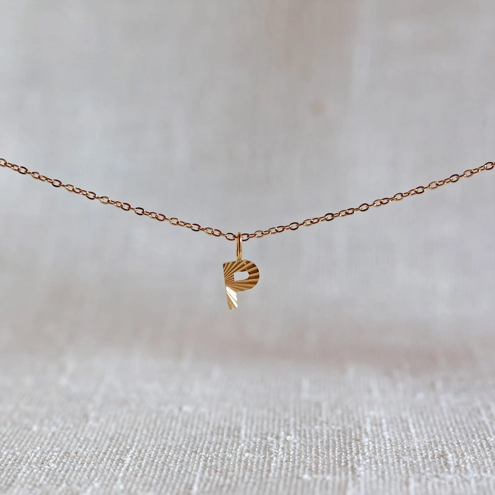Collier "Je suis" avec pendentif lettre  P striée en acier doré, suspendu sur un fond en tissu beige texturé.