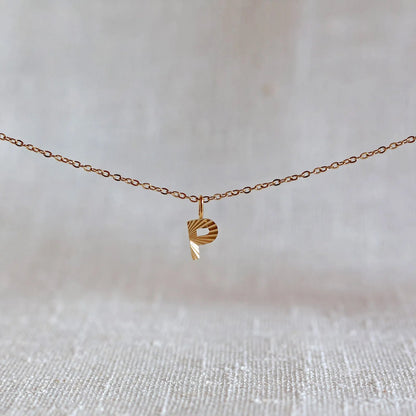 Collier "Je suis" avec pendentif lettre  P striée en acier doré, suspendu sur un fond en tissu beige texturé.