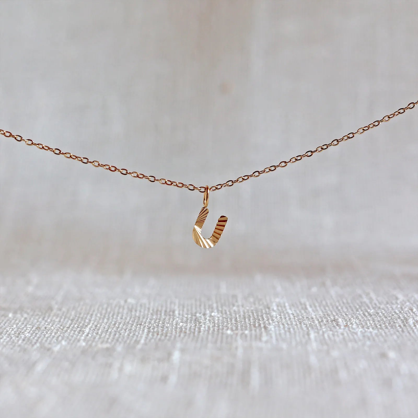 Collier "Je suis" avec pendentif lettre U striée en acier doré, suspendu sur un fond en tissu beige texturé.