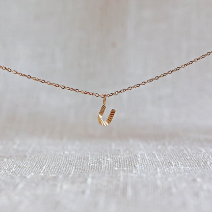 Collier "Je suis" avec pendentif lettre U striée en acier doré, suspendu sur un fond en tissu beige texturé.