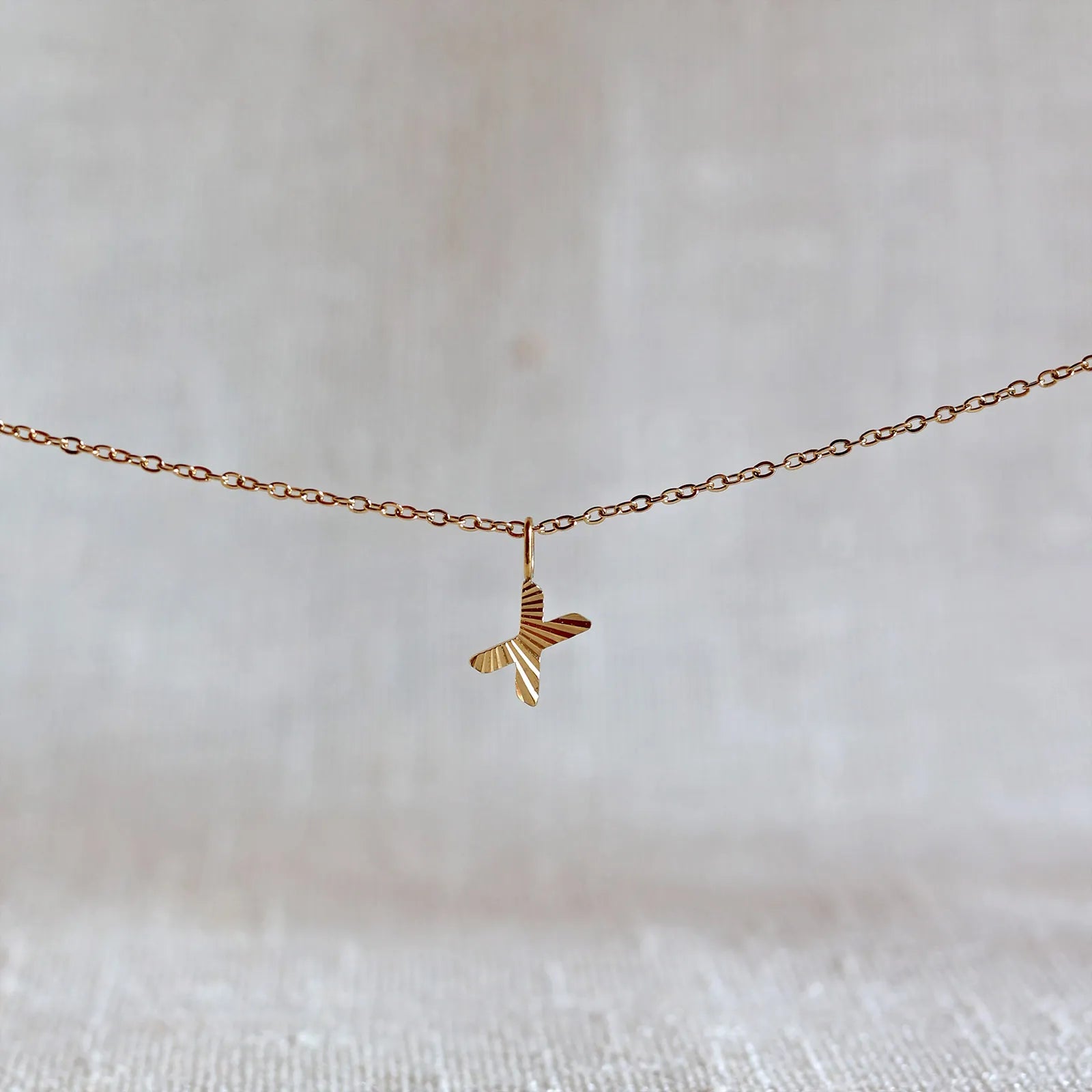 Collier "Je suis" avec pendentif lettre X striée en acier doré, suspendu sur un fond en tissu beige texturé.