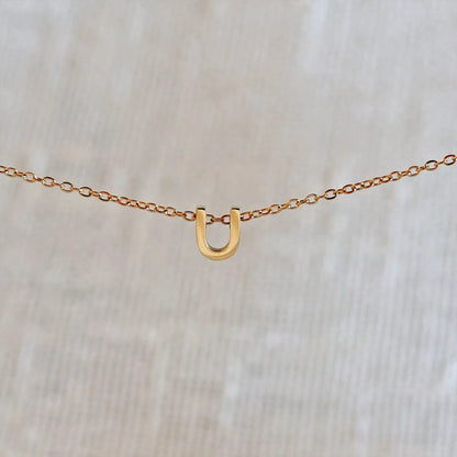 Collier L'instant avec pendentif lettre U doré en acier inoxydable, suspendu au centre d'une maille fine sur fond beige.