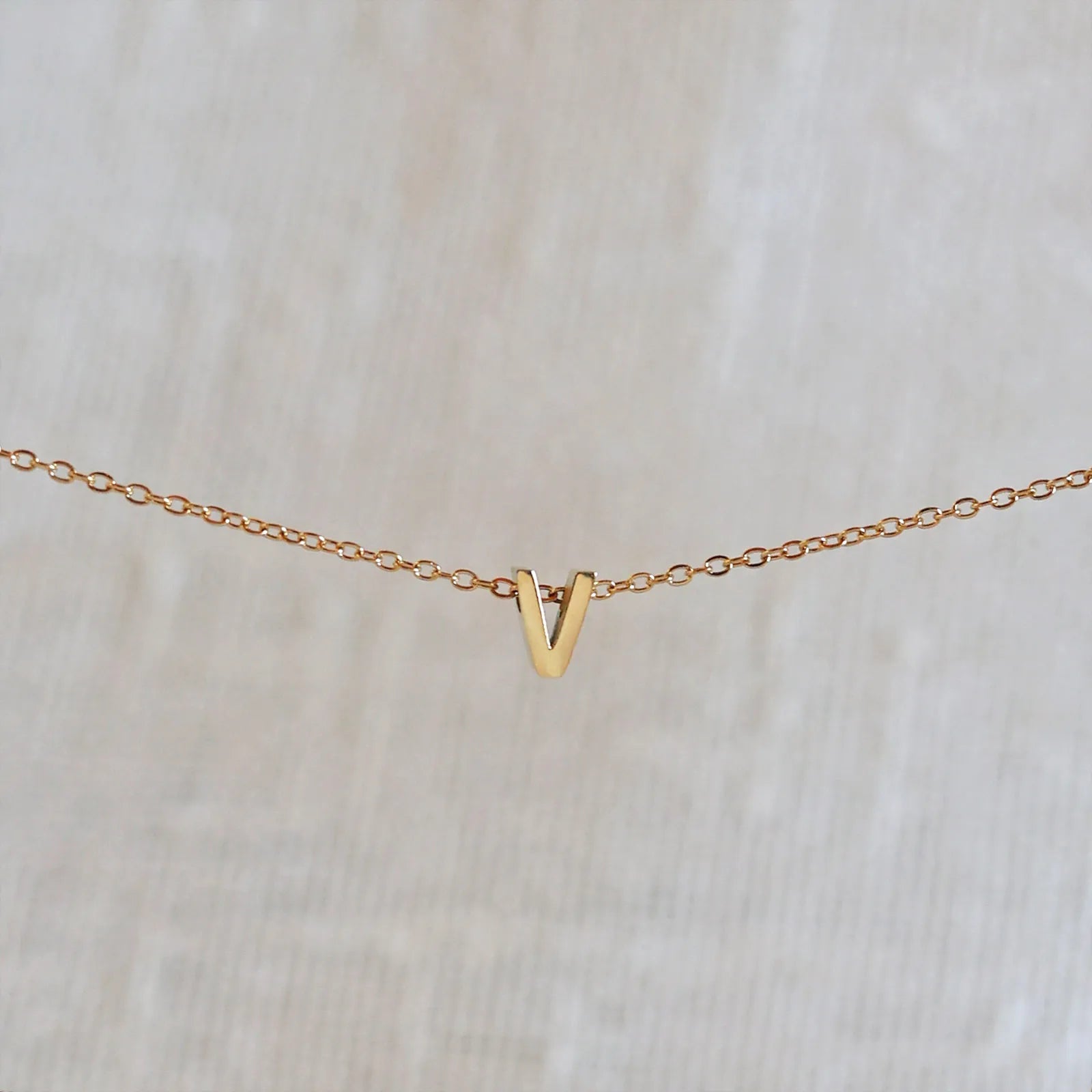Collier L'instant avec pendentif lettre V doré en acier inoxydable, suspendu au centre d'une maille fine sur fond beige.