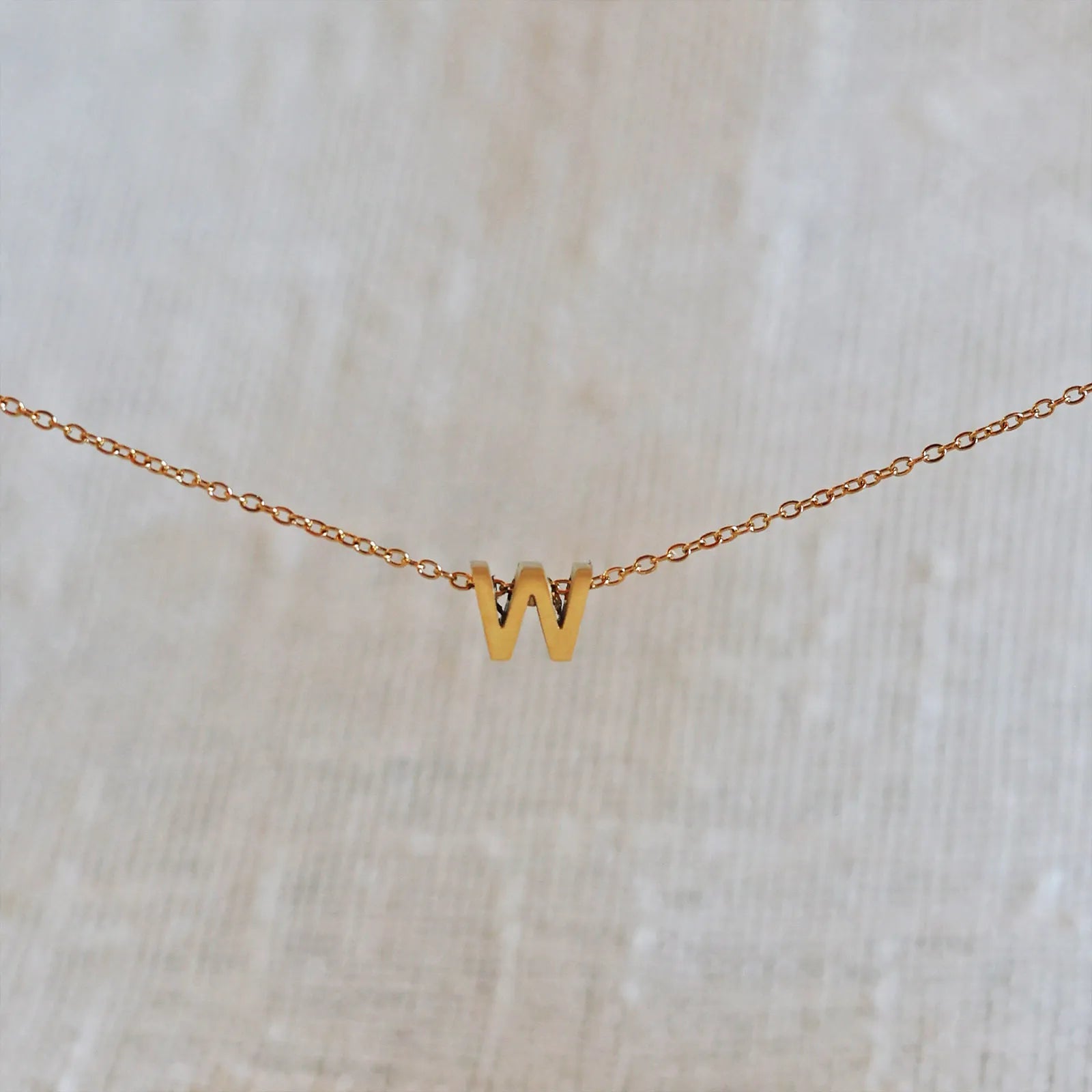 Collier L'instant avec pendentif lettre W doré en acier inoxydable, suspendu au centre d'une maille fine sur fond beige.