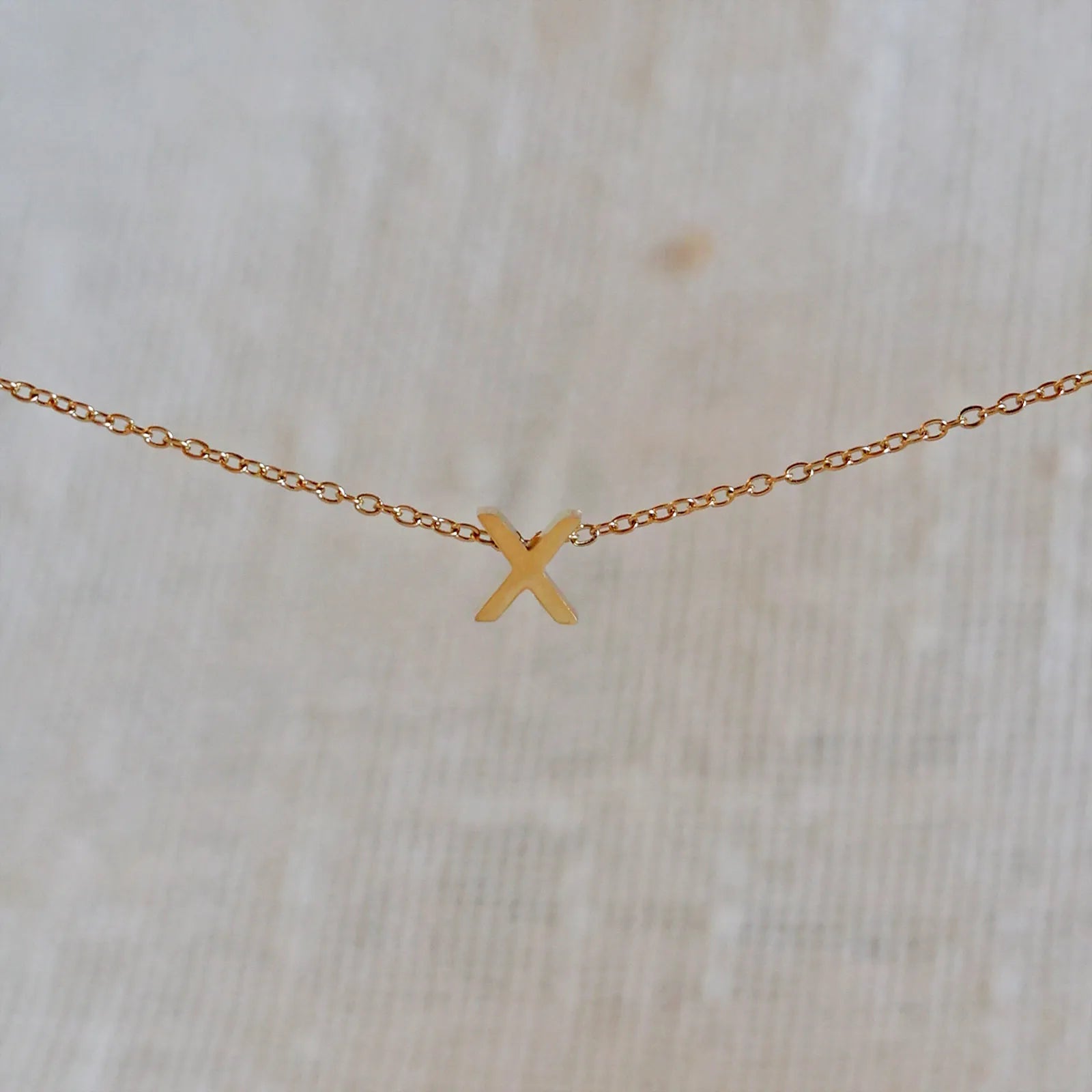 Collier L'instant avec pendentif lettre X doré en acier inoxydable, suspendu au centre d'une maille fine sur fond beige.