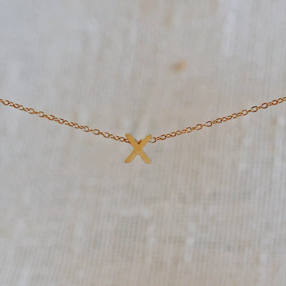 Collier L'instant avec pendentif lettre X doré en acier inoxydable, suspendu au centre d'une maille fine sur fond beige.