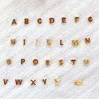 Ensemble de pendentifs lettres dorées en acier inoxydable, soigneusement alignés  sur un tissu beige texturé.