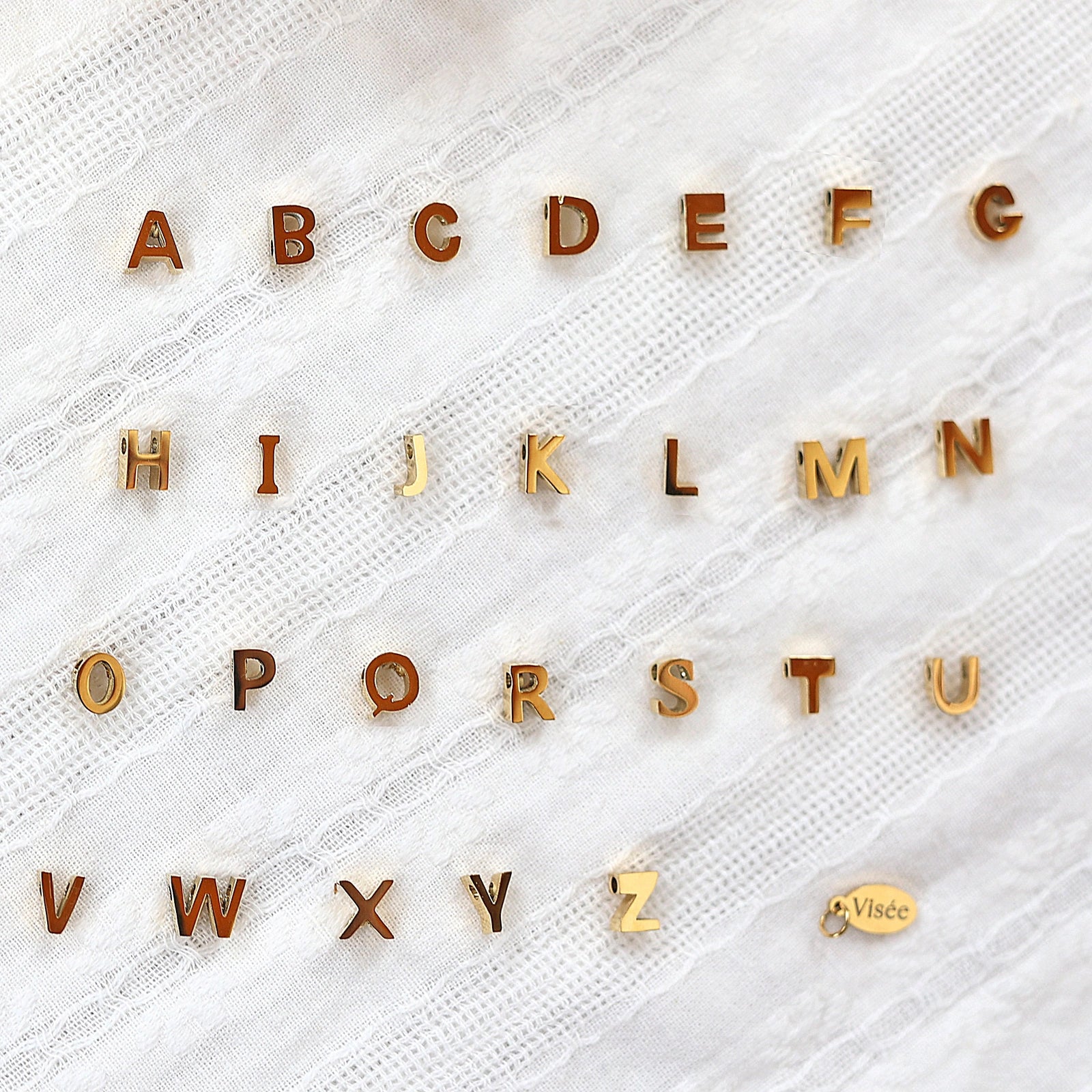 Ensemble de pendentifs lettres dorées en acier inoxydable, soigneusement alignés  sur un tissu beige texturé.