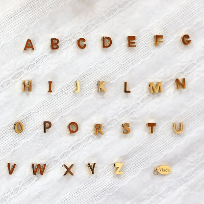 Ensemble de pendentifs lettres dorées en acier inoxydable, soigneusement alignés  sur un tissu beige texturé.