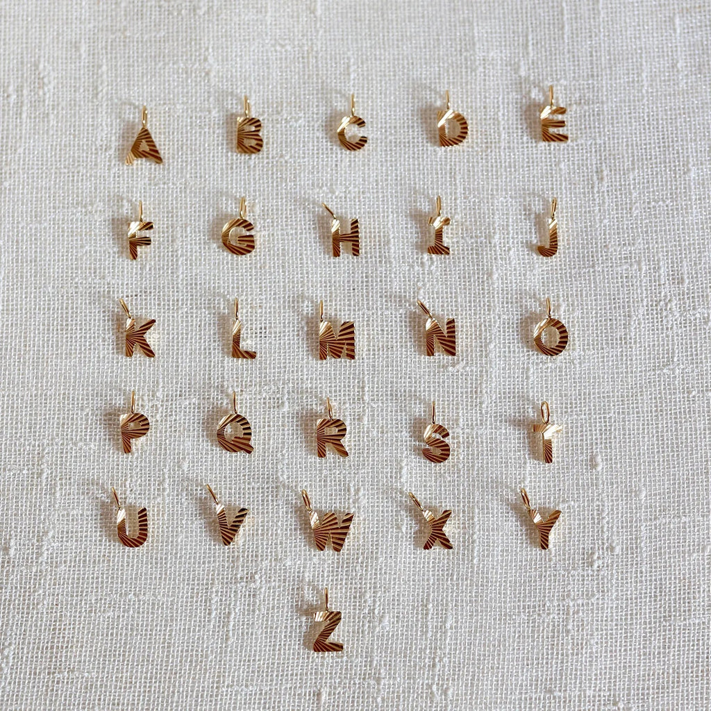 Ensemble de pendentifs lettres dorées en acier inoxydable, soigneusement alignés  sur un tissu beige texturé.