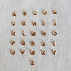 Ensemble de pendentifs lettres dorées en acier inoxydable, soigneusement alignés  sur un tissu beige texturé.