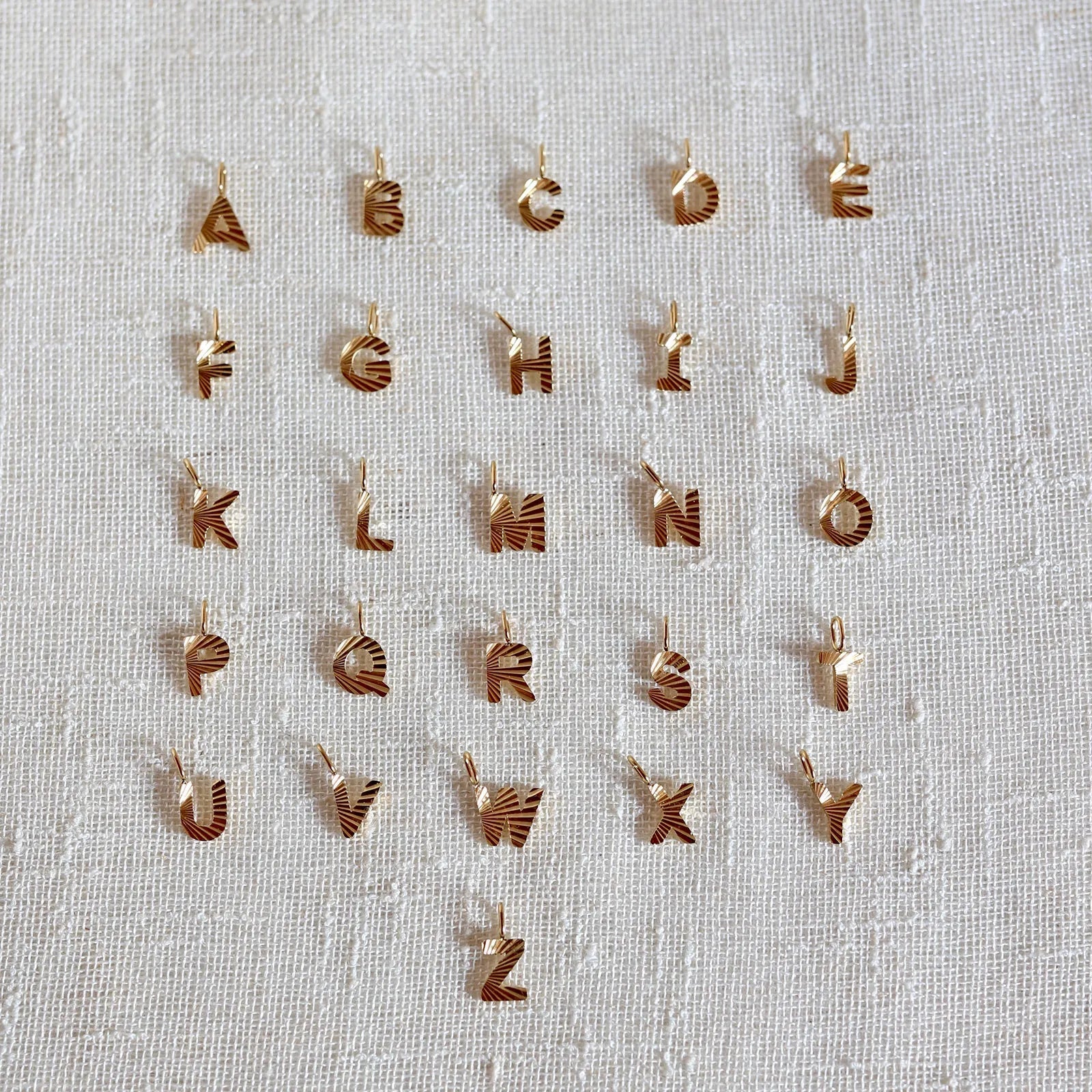 Ensemble de pendentifs lettres dorées en acier inoxydable, soigneusement alignés  sur un tissu beige texturé.