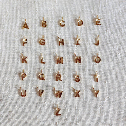 Ensemble de pendentifs lettres dorées en acier inoxydable, soigneusement alignés  sur un tissu beige texturé.