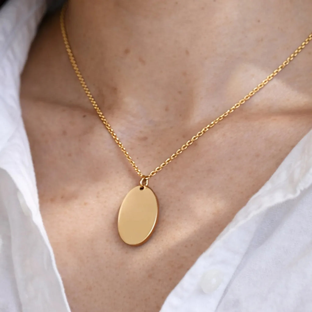 Zoom sur le collier "Tendresse", pendentif ovale personnalisable en acier inoxydable doré, porté autour du cou, avec chemise blanche