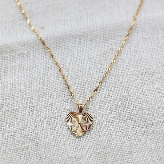 Collier A coeur ouvert avec pendentif cœur strié en acier doré personnalisé, chaîne torsadée, posé sur un tissu beige texturé.