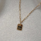 Collier "Ma vérité" avec pendentif rectangulaire personnalisable en acier doré, présenté sur un fond beige texturé.