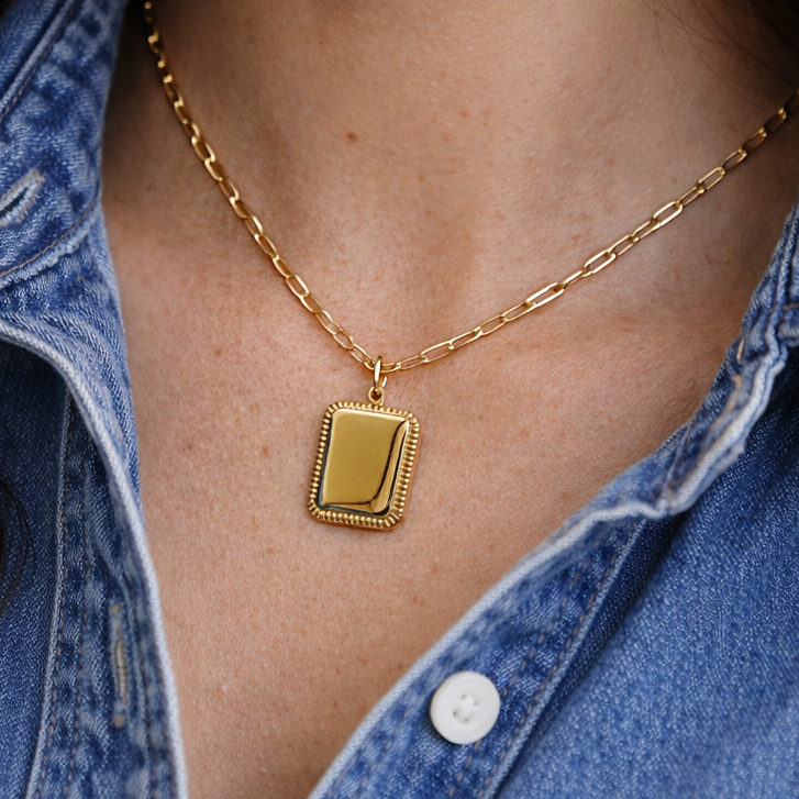 Collier Trésor avec pendentif rectangulaire personnalisable en acier doré, porté autour du cou d'une jeune femme qui porte une chemise jeans bleu.