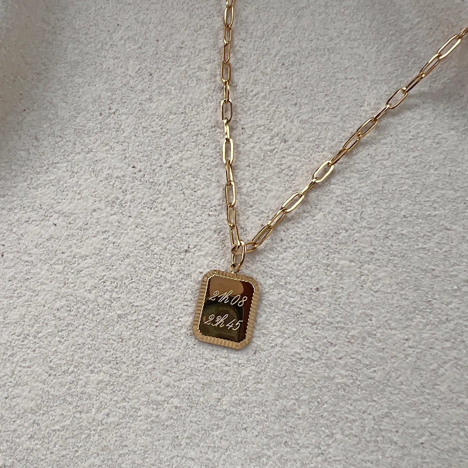 Collier Trésor avec pendentif rectangulaire personnalisé avec heure de naissance en acier doré, présenté sur un fond beige texturé.