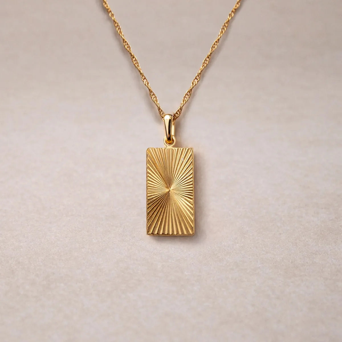 Collier Eclat  en acier doré avec pendentif rectangulaire gravé de rayons, personnalisable au verso, vue de face sur support beige texturé.